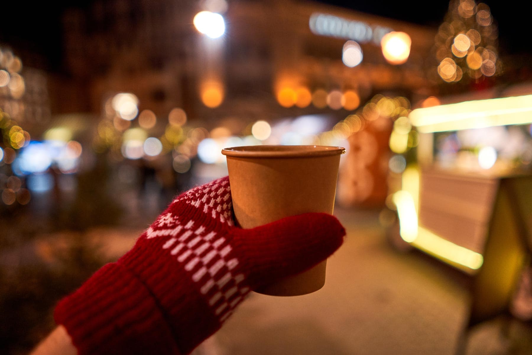 Nahaufnahme von einer behandschuhten Hand, die ein Heißgetränk hält, im Hintergrund ist ein Weihnachtsmarkt in Sachsen-Anhalt zu sehen